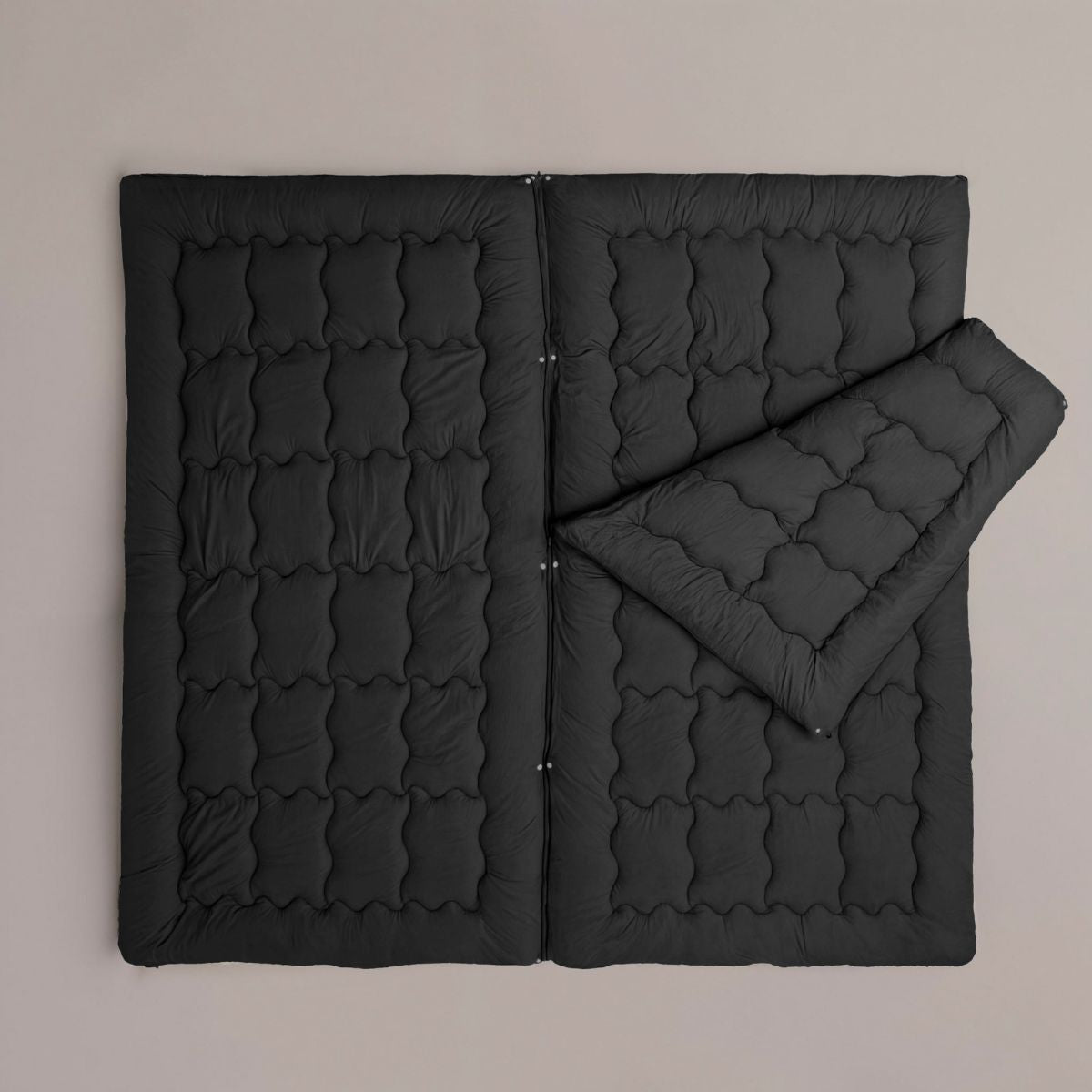Le Zelesta Flexibed - Noir, une couette matelassée ajustable noire, se pose à plat sur une surface neutre avec un côté replié, offrant confort et chaleur adaptable pour une température idéale.