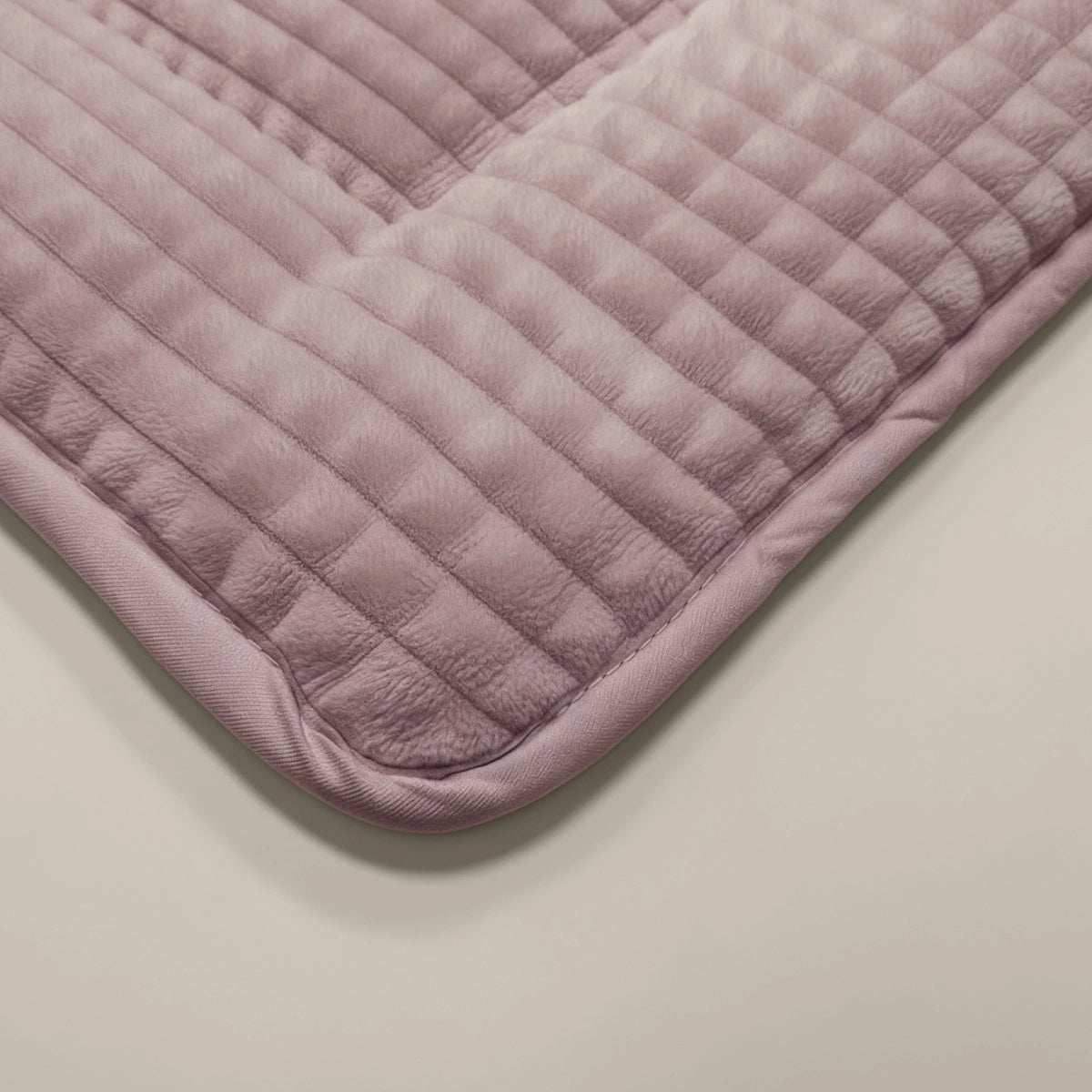 Gros plan sur le tapis Zelesta Teddy Check - Vieux Rose, composé d'un tissu matelassé violet clair à la texture douce et pelucheuse, aux coins arrondis et aux détails cousus, sur un fond neutre.