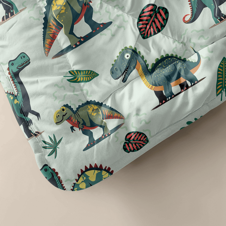 Le Kiddybed - Dino de Zelesta est une couverture de couleur claire avec des dinosaures en dessin animé et des feuilles vertes, conçue pour les enfants et facile à nettoyer grâce à sa conception lavable.